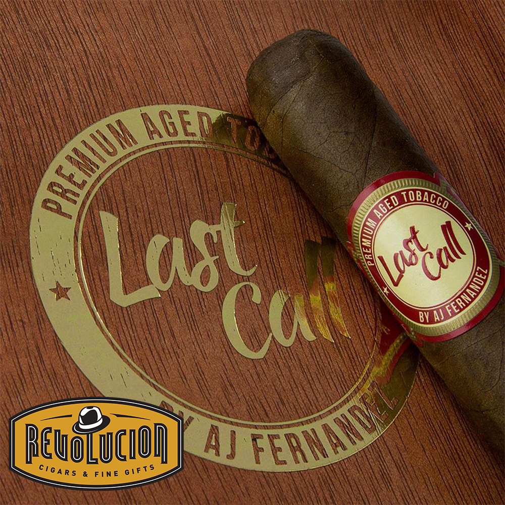 AJ Fernandez Last Call Geniales Habano cigar placed diagonally on a wooden surface, with the cigar's white and red band and the 'Last Call' logo prominently displayed in the background