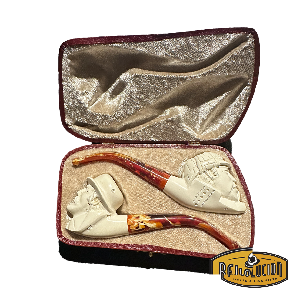 A set of two Meerschaum pipes in a velvet-lined case. Both pipes have cream-colored bowls with intricate carvings and red marbled stems. One pipe features a carved face of a man with a hat, and the other has a different face carving.