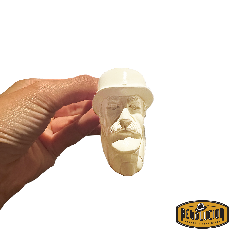 A hand holding a Meerschaum pipe with a carved face of a man with a mustache, wearing a hat. The pipe is cream-colored and intricately detailed.