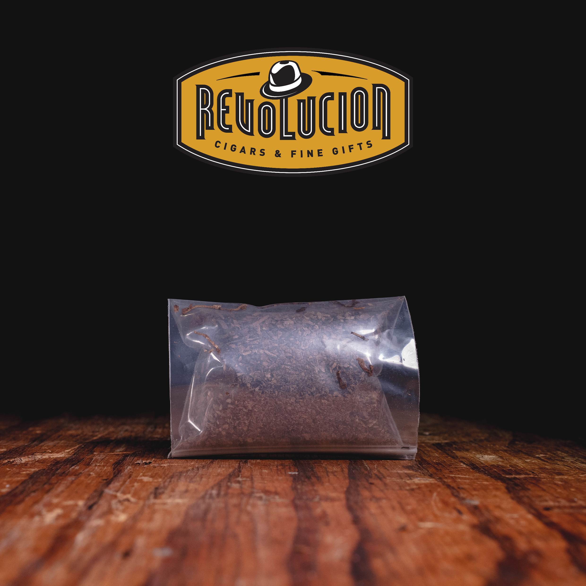 A sealed bag of finely cut tobacco placed on a wooden surface, with the "Revolucion Cigars & Fine Gifts" logo above.
