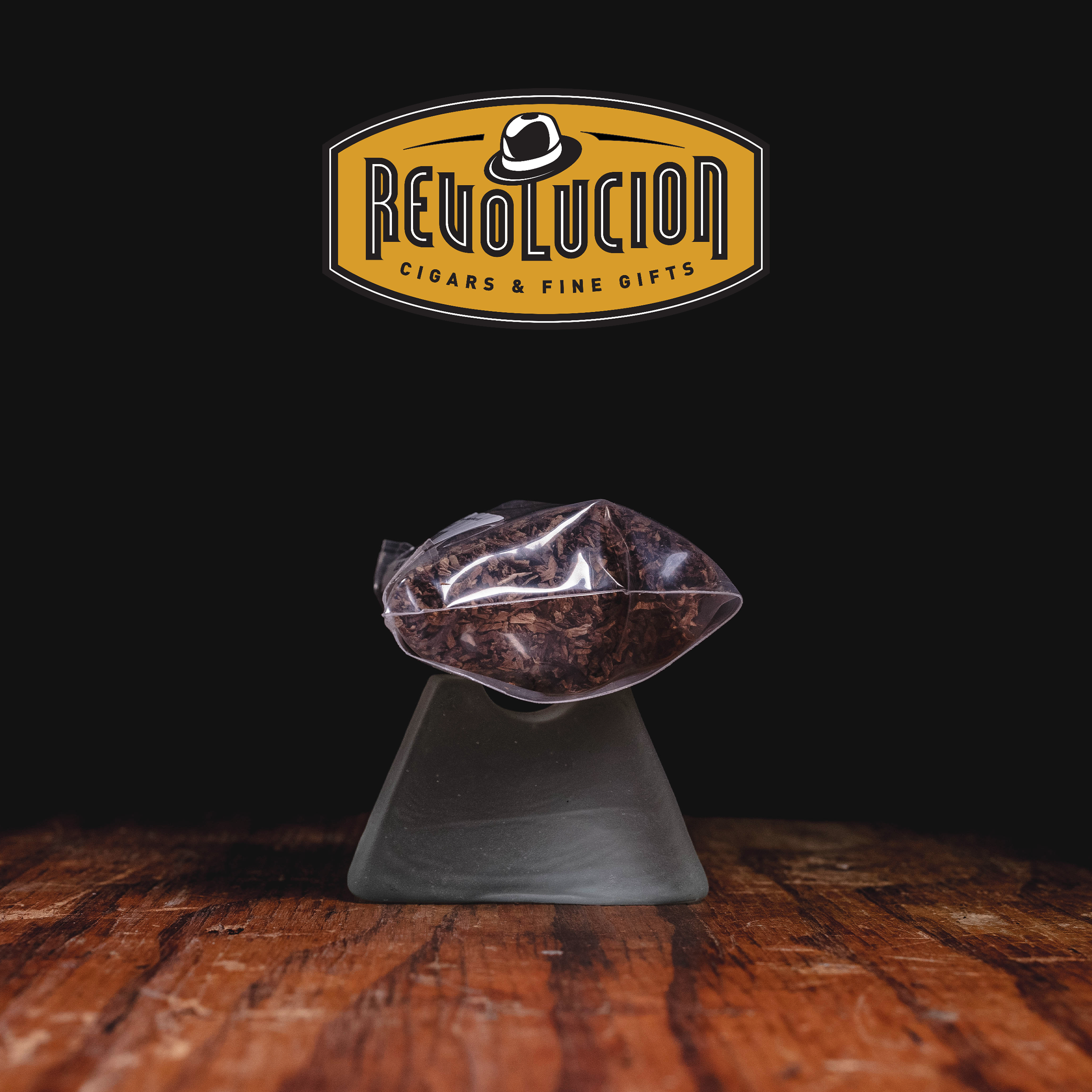 A bag of loose tobacco resting on a glass stand, set against a dark background with the "Revolucion Cigars & Fine Gifts" logo above.