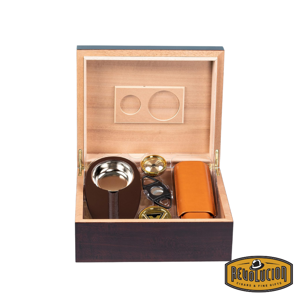 An open wooden cigar humidor with accessories neatly arranged inside, including an ashtray, a cigar case, a cutter, a hygrometer, and a humidifier. The interior is lined with cedar, featuring slots for humidification elements. The Revolucion Cigars & Fine Gifts logo is in the bottom right corner.