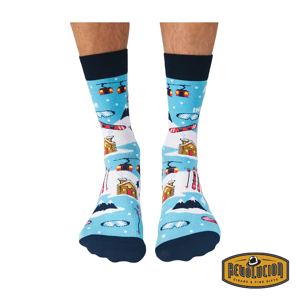 Front view of blue 'Après Ski Instructor' socks featuring ski lifts, cabins, skis, and snowy mountain graphics. Socks are blue with black cuffs and toes, branded with the Revolucion Cigars & Fine Gifts logo.