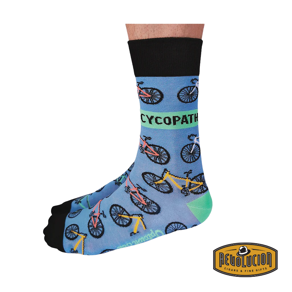 Side view of blue socks featuring colorful bicycle graphics and the word 'CYCOPATH' in bold green letters. The socks have black cuffs and toes and are branded with the Revolucion Cigars & Fine Gifts logo.