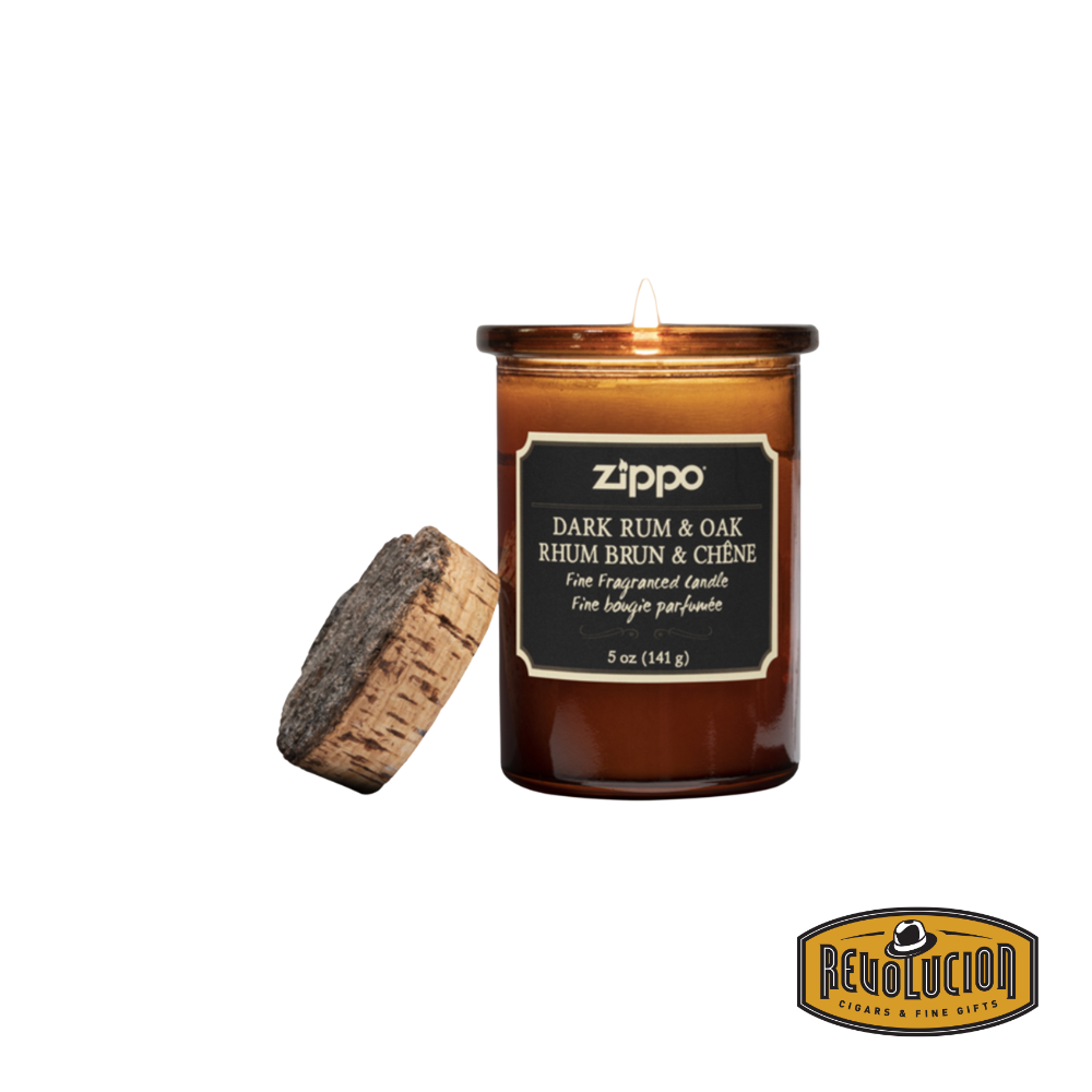 Zippo Dark Rum & Oak Candle in an amber glass jar with a cork lid, glowing warmly. The label highlights the fragrance name and weight in both English and French.