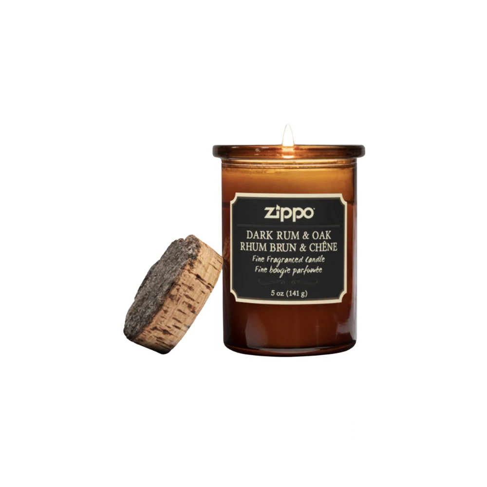 Zippo Dark Rum & Oak Candle in an amber glass jar with a cork lid, glowing warmly. The label highlights the fragrance name and weight in both English and French.