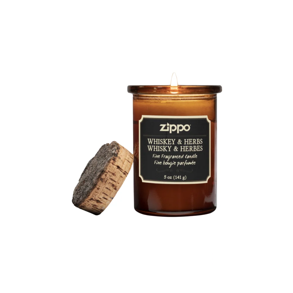 Zippo Dark Whiskey & Herbs Candle in an amber glass jar with a cork lid, emitting a warm glow. The label showcases the fragrance name and weight in both English and French.