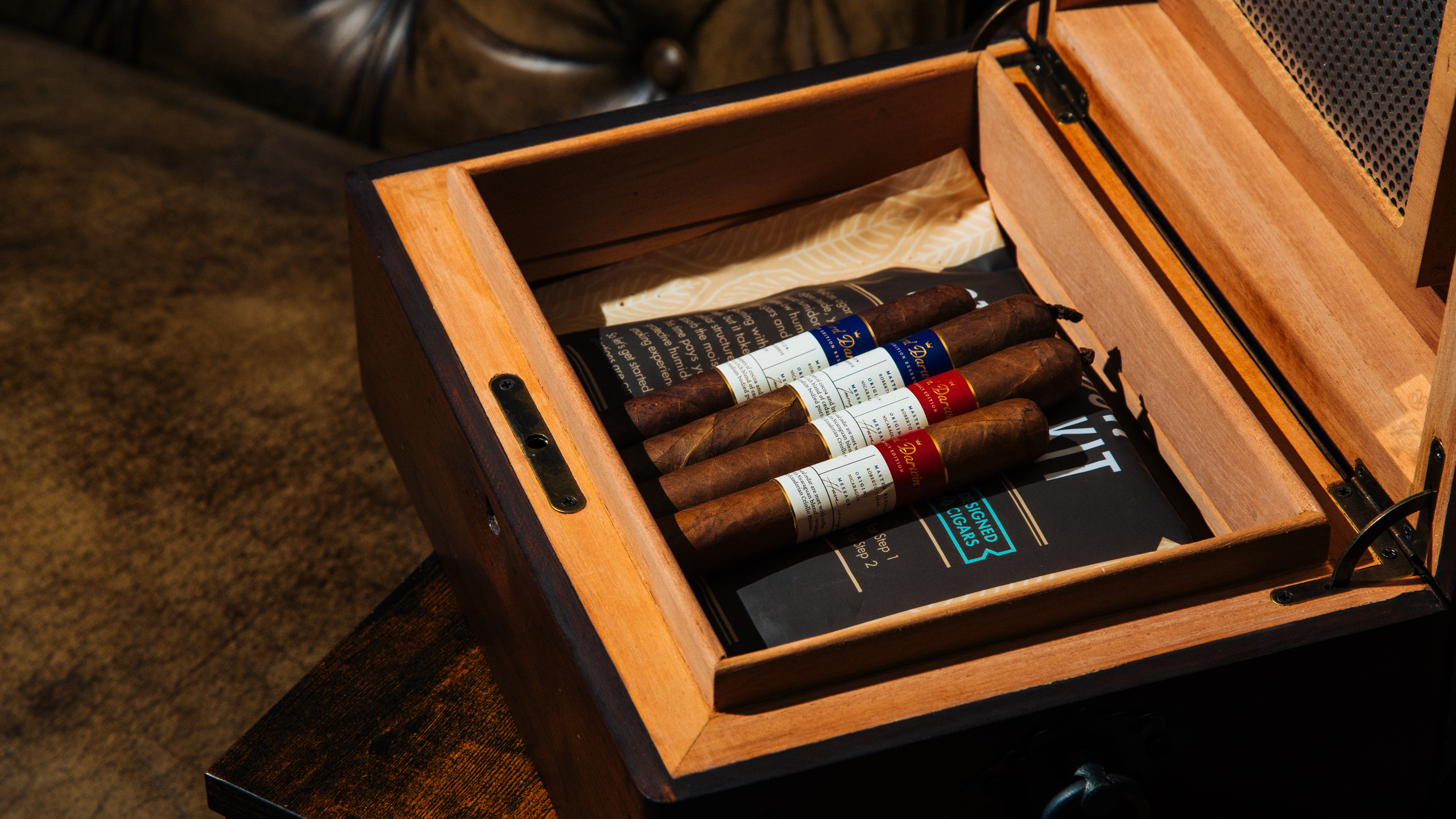 Open wooden humidor with cigars and humidor accessories on a leather couch surface.