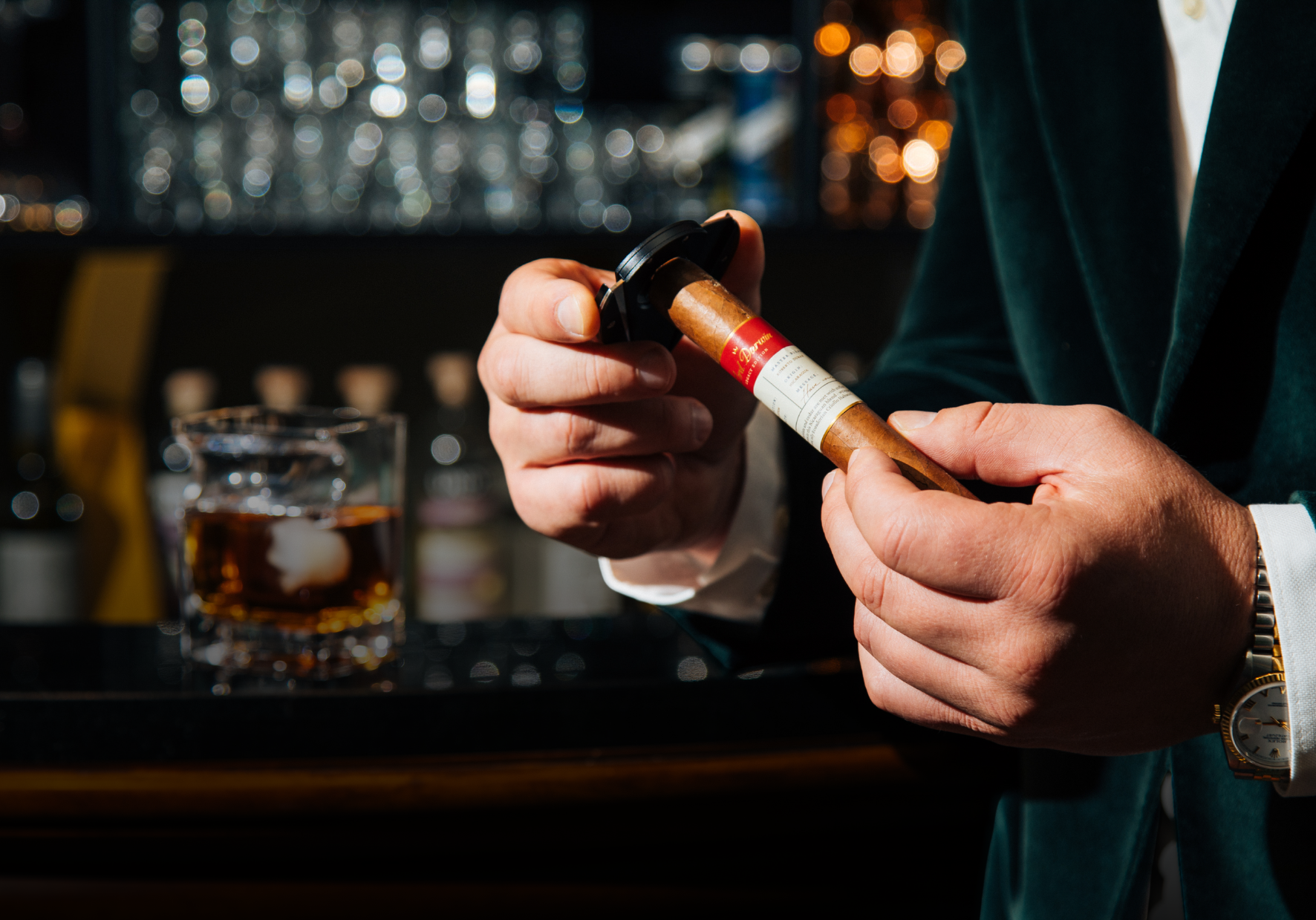Person holding a revolucion lord darwin exclusive premium cigar with a glass of whiskey in a bar setting