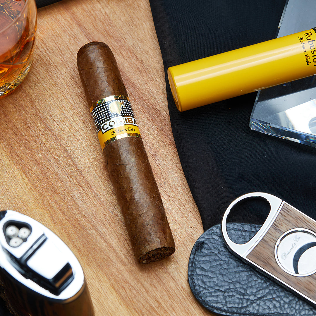 Cohiba Robusto Cuban cigar with a golden yellow band, placed on a wooden surface, accompanied by a black cloth, a cigar cutter, a lighter, and a yellow Cohiba tubo.