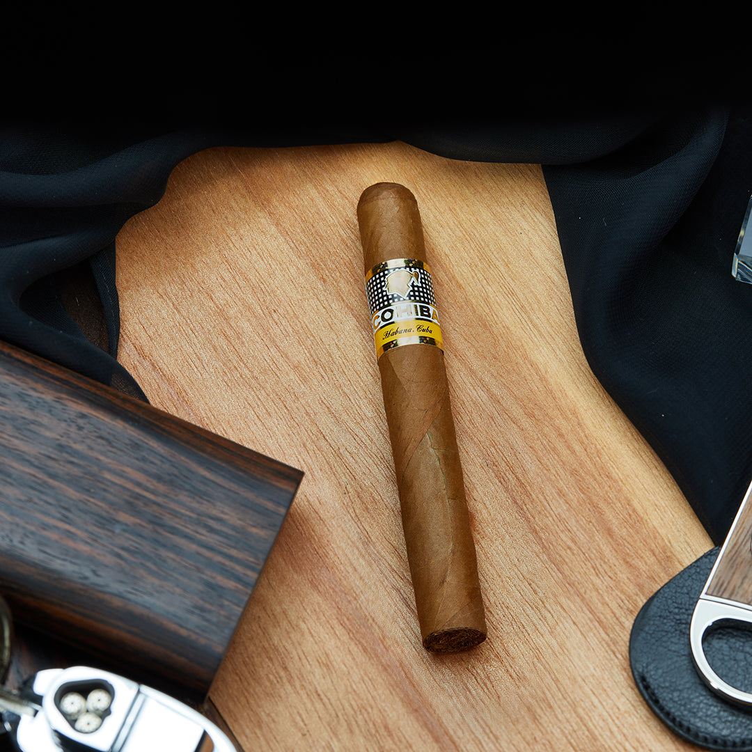 Cohiba Siglo II Cuban cigar with a golden yellow band, resting on a wooden surface, surrounded by a black cloth, a cigar cutter, and wooden accessories.