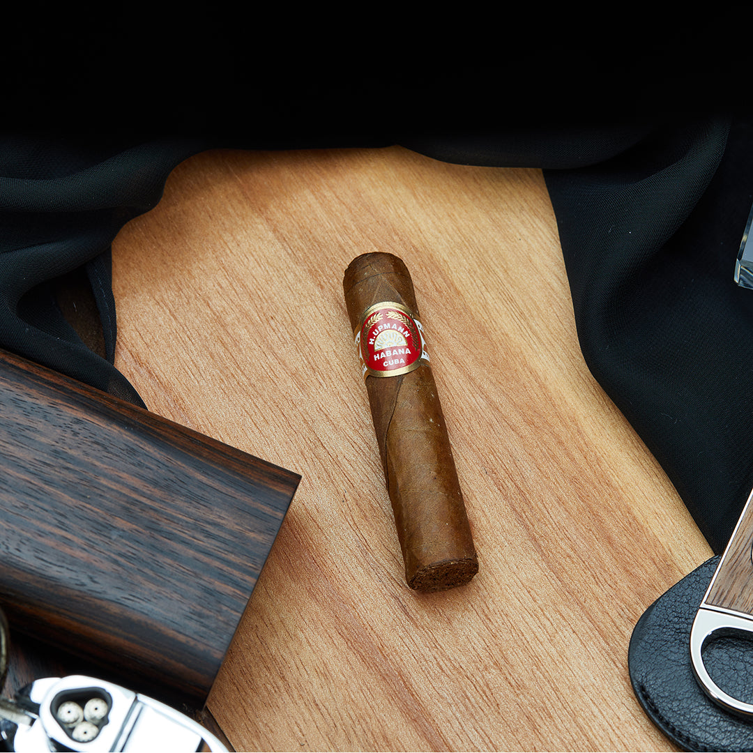 H. Upmann Half Corona Cuban cigar with a red and gold band, resting on a wooden surface with a cutter, lighter, and a black cloth in the background.