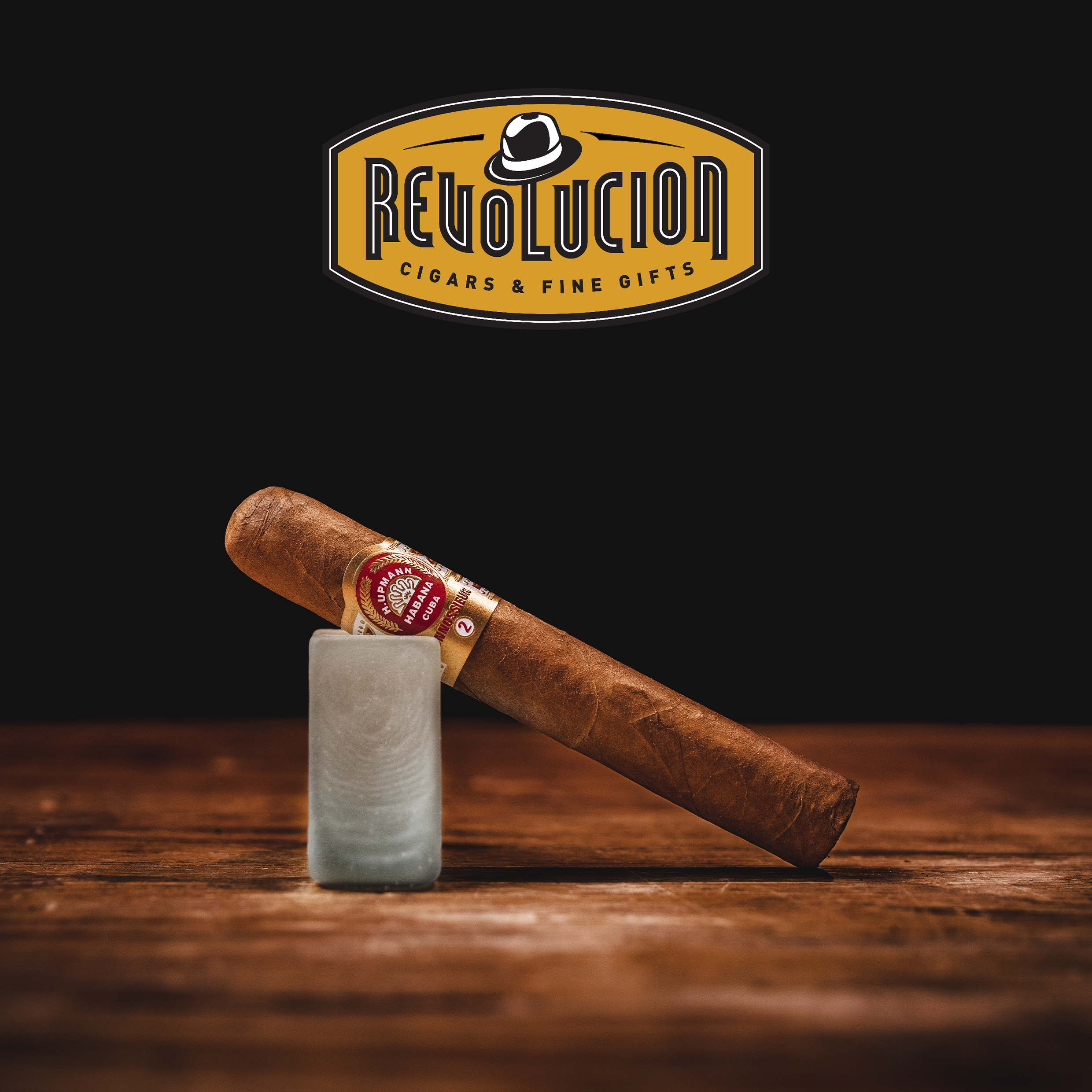 H. Upmann Connossieur No. 2 Cuban cigar resting on a glass stand with the Revolucion Cigars & Fine Gifts logo in the background.
