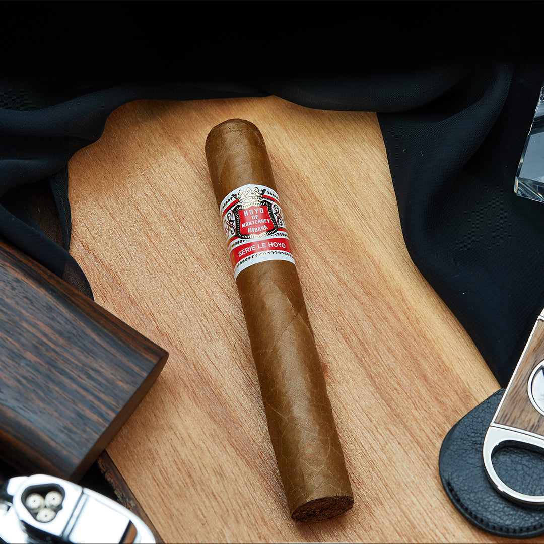 Hoyo de Monterrey San Juan cigar with a red and white band, placed on a wooden surface alongside a cutter, a lighter, and a glass of whiskey.