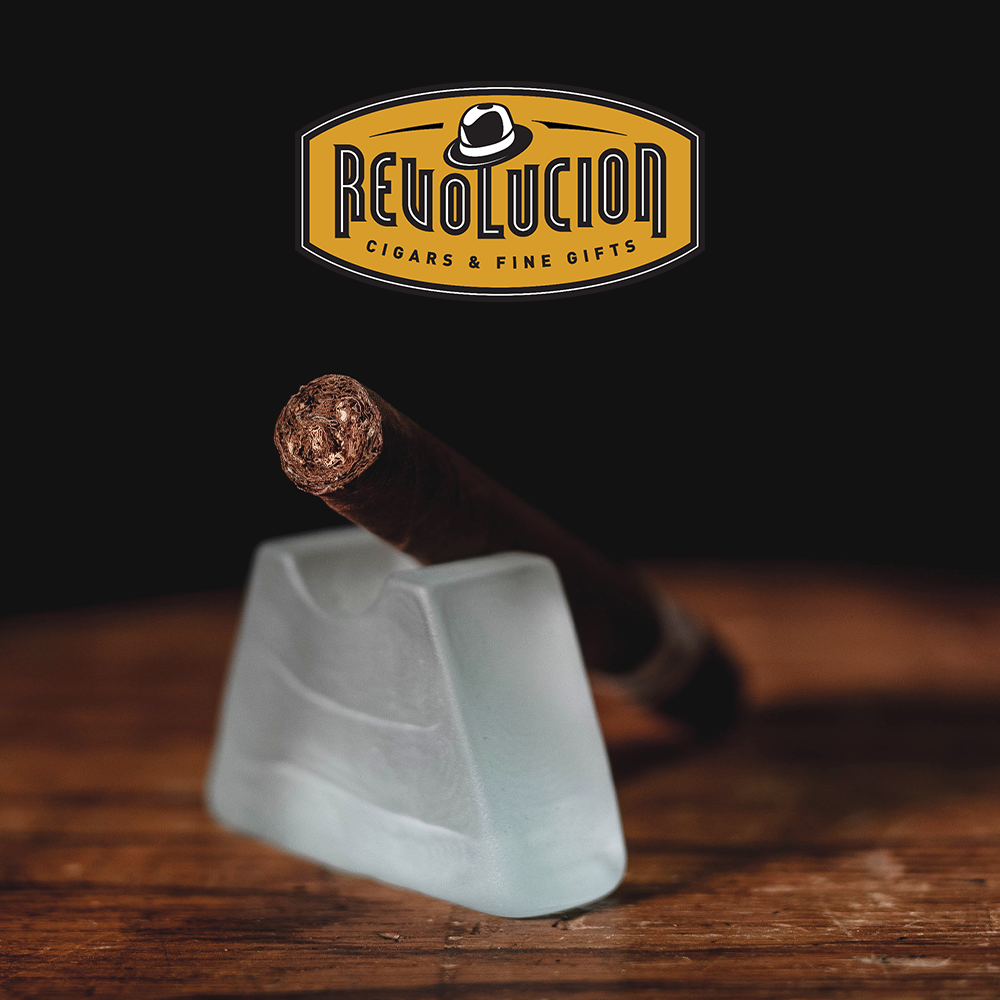 Jose L. Piedra Cremas Cuban cigar resting on a wooden surface with the Revolucion Cigars & Fine Gifts logo in the background.