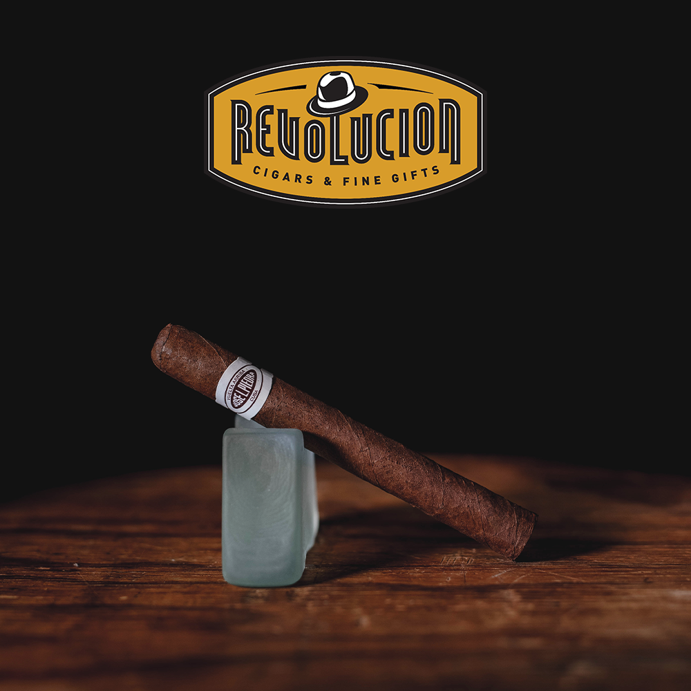Jose L. Piedra Cremas Cuban cigar resting on a wooden surface with the Revolucion Cigars & Fine Gifts logo in the background.