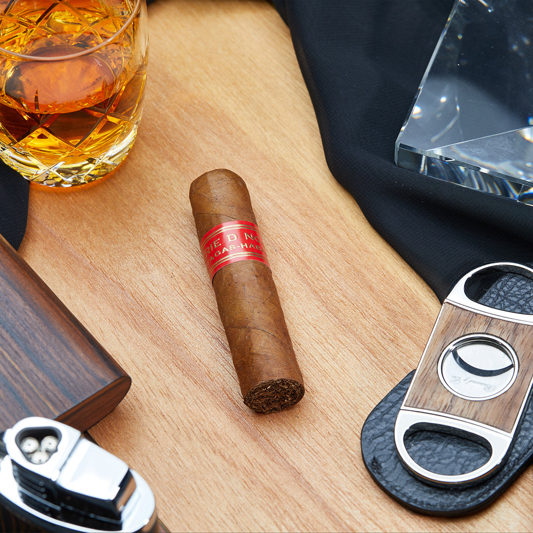 Partagás Serie D No. 6 Cuban cigar with a red band, placed on a wooden surface alongside a whiskey glass, a cigar cutter, and other accessories.