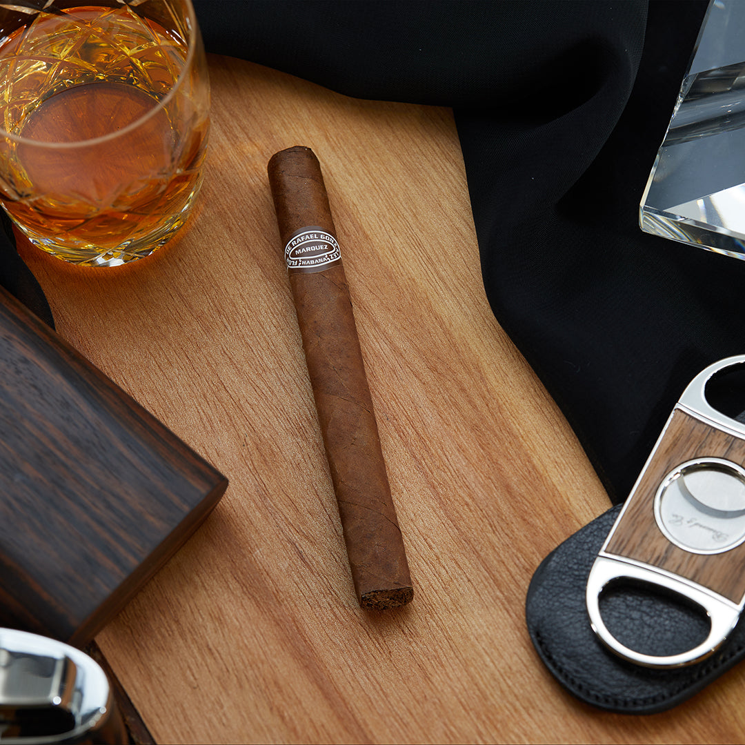 Rafael González Panetela Extra Cuban cigar with a brown wrapper and an elegant band, resting on a wooden surface alongside a whiskey glass, cutter, and other cigar accessories.