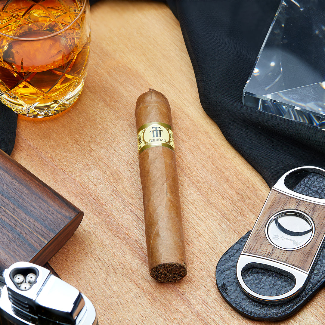 Trinidad Media Luna Cuban cigar placed on a wooden surface, accompanied by a whiskey glass, cigar cutter, and lighter, showcasing its golden band and signature pigtail cap.
