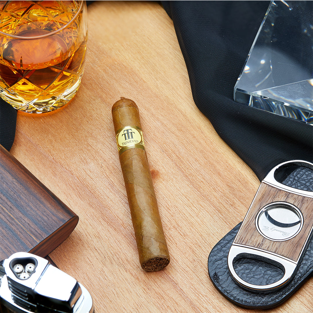 Trinidad Reyes Cuban cigar on a wooden surface, surrounded by a whiskey glass, a cigar cutter, and a lighter, featuring its golden band and classic pigtail cap.