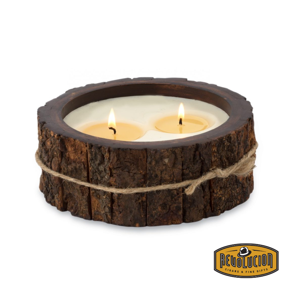 Rustic handmade candle in a round tree bark pot, featuring two lit wicks, accented with a decorative jute cord tied around the natural bark exterior. Includes the Revolucion Cigars & Fine Gifts logo.