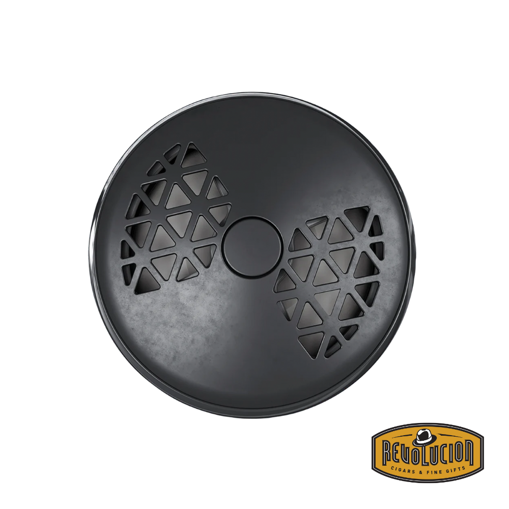 The photo shows the front of the Maison Berger Smart Car Diffuser, featuring a sleek and modern design in tones of black and gunmetal. The device has small triangular openings that form a geometric pattern reminiscent of a bowtie. The device is turned to the left, indicating a lower fragrance intensity.