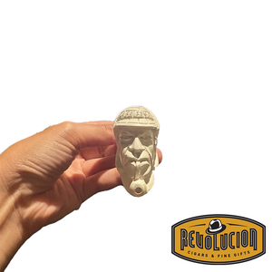 A hand holding a Meerschaum pipe with a carved face of a man wearing a helmet. The pipe is cream-colored and showcases intricate facial details from the front view