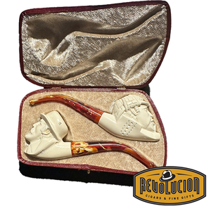 A set of two Meerschaum pipes in a velvet-lined case. Both pipes have cream-colored bowls with intricate carvings and red marbled stems. One pipe features a carved face of a man with a hat, and the other has a different face carving