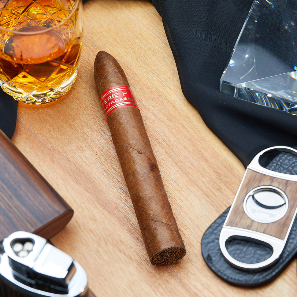 Partagás Serie P No.2 cigar resting on a wooden surface, surrounded by a glass of whiskey, a cigar cutter, and a lighter – showcasing a classic cigar lounge vibe.