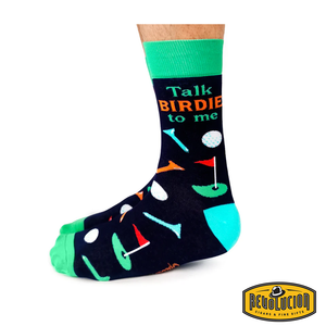 Side view of black socks with golf-themed graphics and the phrase 'Talk Birdie to Me' in orange and green letters. The socks have green cuffs, green toes, and turquoise heels, branded with the Revolucion Cigars & Fine Gifts logo.