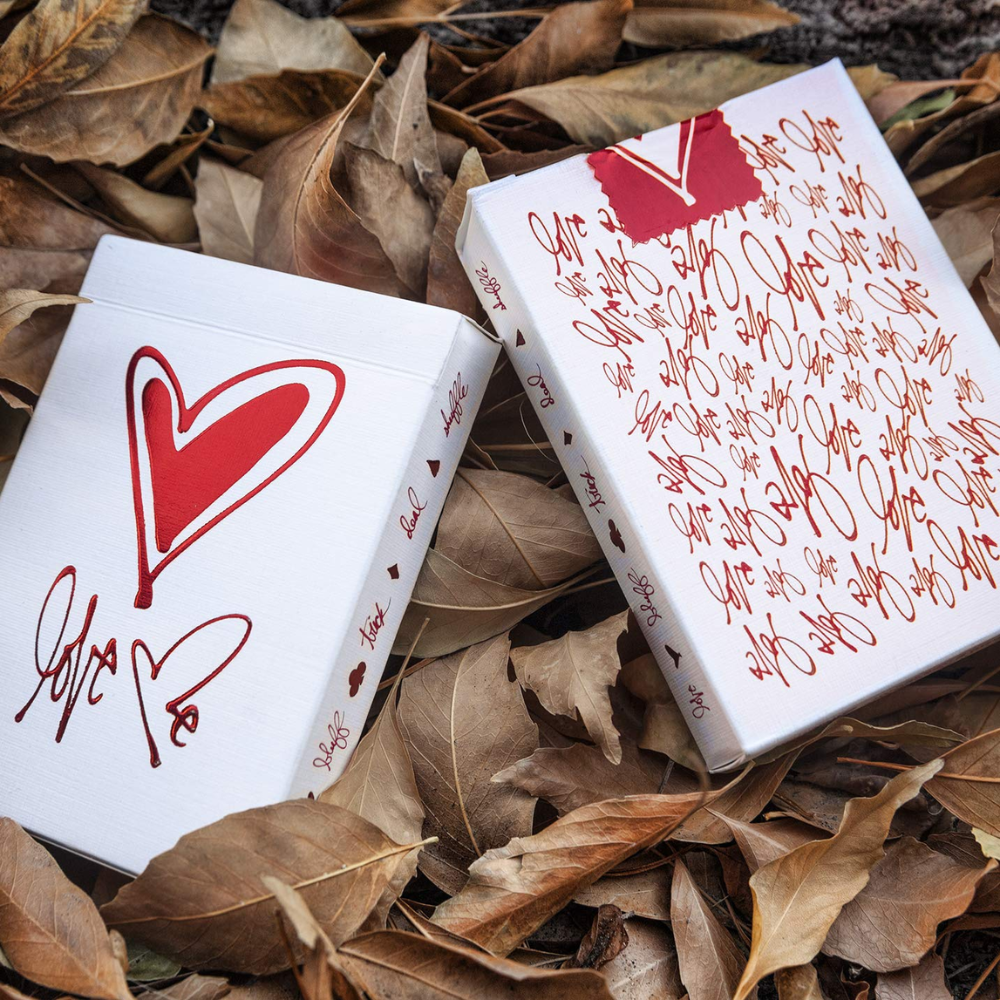 Theory 11 Love Me Playing Cards box showing front and back design, placed in a romantic setting with leaves in the background.