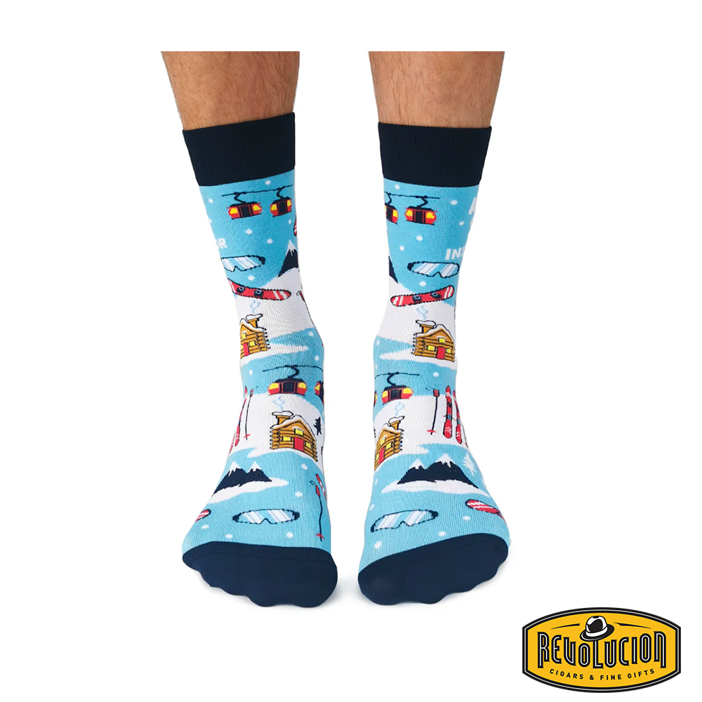 Front view of blue &#39;Après Ski Instructor&#39; socks featuring ski lifts, cabins, skis, and snowy mountain graphics. Socks are blue with black cuffs and toes, branded with the Revolucion Cigars &amp; Fine Gifts logo.