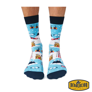 Front view of blue 'Après Ski Instructor' socks featuring ski lifts, cabins, skis, and snowy mountain graphics. Socks are blue with black cuffs and toes, branded with the Revolucion Cigars & Fine Gifts logo.