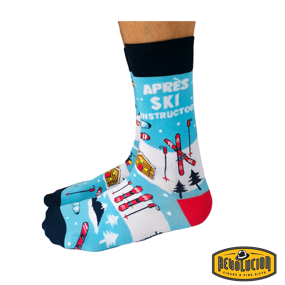 Front view of blue 'Après Ski Instructor' socks featuring ski lifts, cabins, skis, and snowy mountain graphics. Socks are blue with black cuffs and toes, branded with the Revolucion Cigars & Fine Gifts logo.