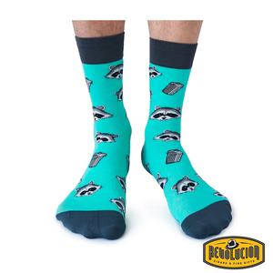 Front view of teal socks featuring playful raccoon and trash can graphics, with dark gray cuffs and toes. The socks are branded with the Revolucion Cigars & Fine Gifts logo