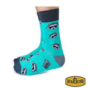 Side view of teal socks featuring playful raccoon and trash can graphics, with dark gray cuffs, heels, and toes. The socks are branded with the Revolucion Cigars & Fine Gifts logo