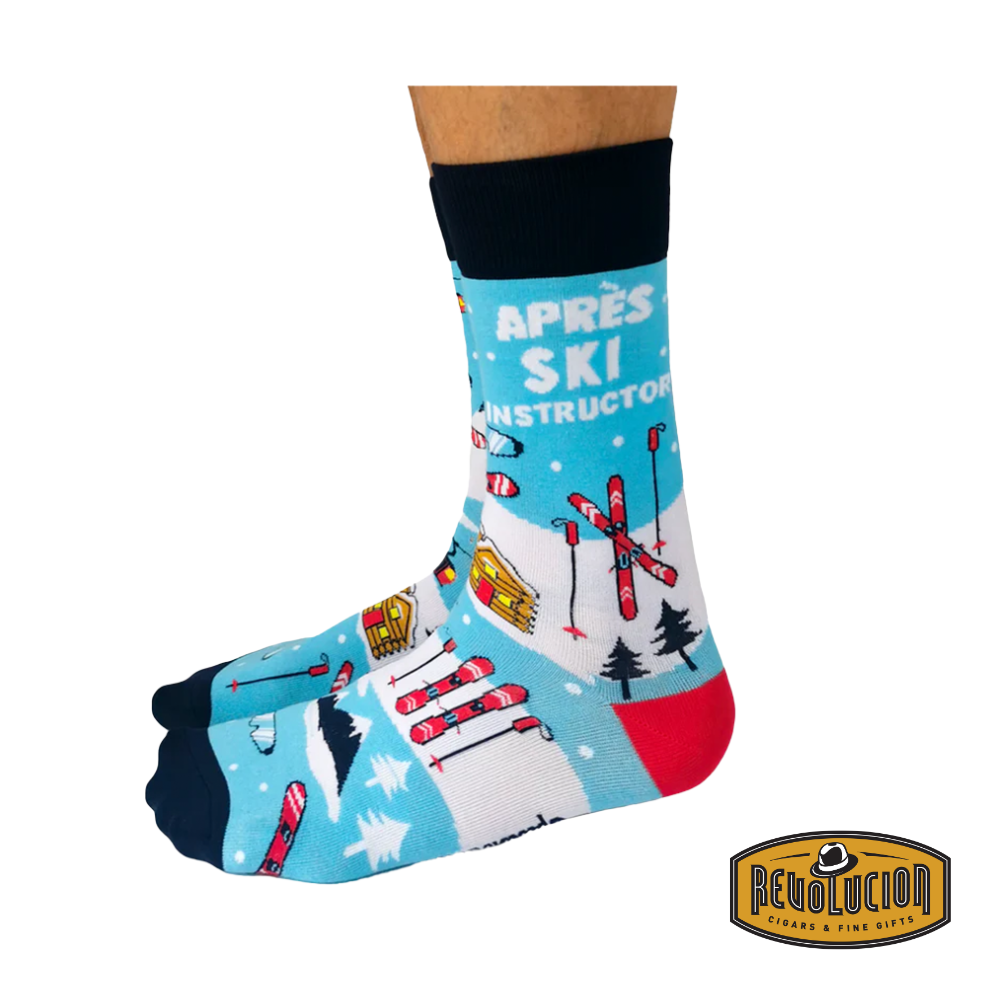 Side view of blue 'Après Ski Instructor' socks with detailed graphics of ski lifts, cabins, skis, and snowy mountains. Socks are blue with black cuffs and toes, branded with the Revolucion Cigars & Fine Gifts logo.