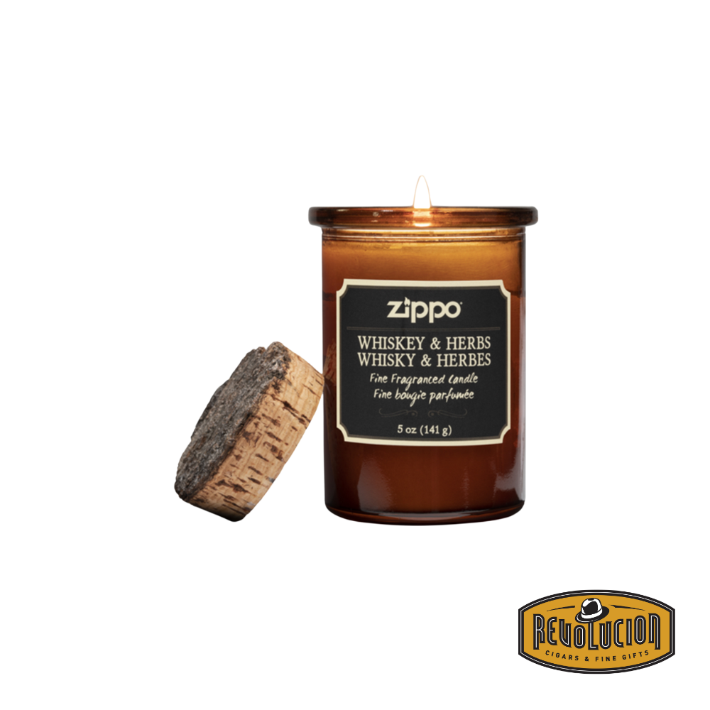 Zippo Dark Whiskey & Herbs Candle in an amber glass jar with a cork lid, emitting a warm glow. The label showcases the fragrance name and weight in both English and French.