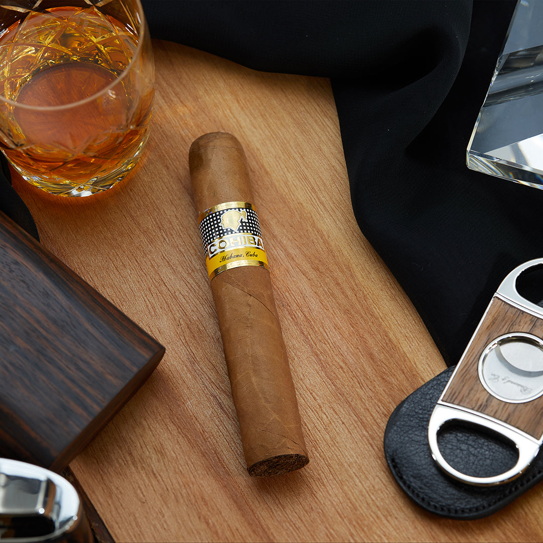 Cohiba Robusto Cuban cigar with a golden yellow band, placed on a wooden surface, accompanied by a black cloth, a cigar cutter, a lighter, a cigar case and a whisky glass.