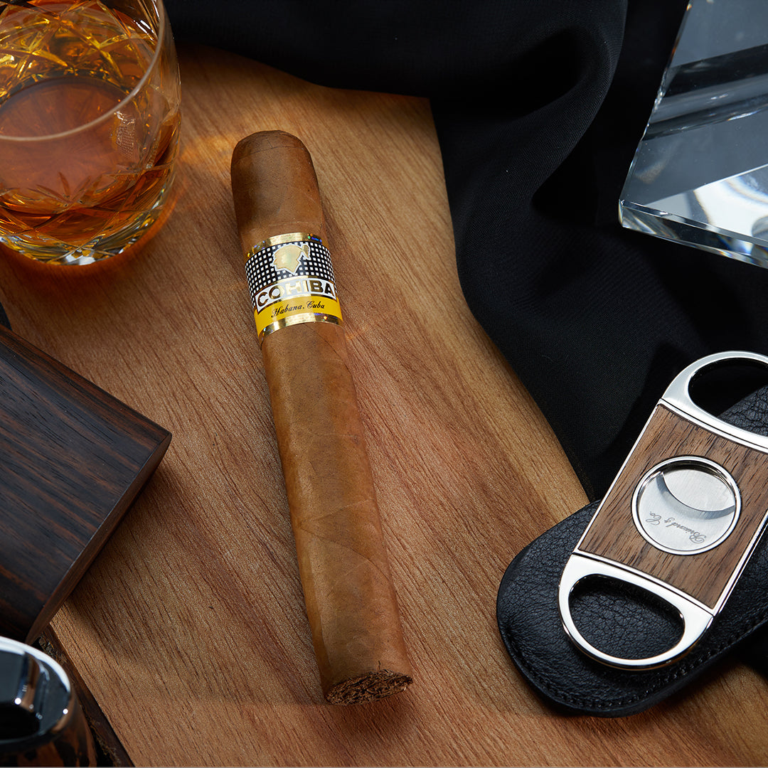 Cohiba Siglo VI Cuban cigar with a golden yellow band, placed on a wooden surface alongside a glass of whiskey, a cigar cutter, and a black cloth.
