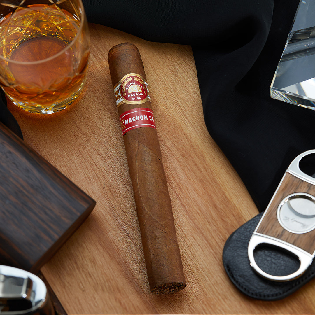 H. Upmann Magnum 50 Cuban cigar with a red and gold band, placed on a wooden surface alongside a cutter, a lighter, and a glass of whiskey.