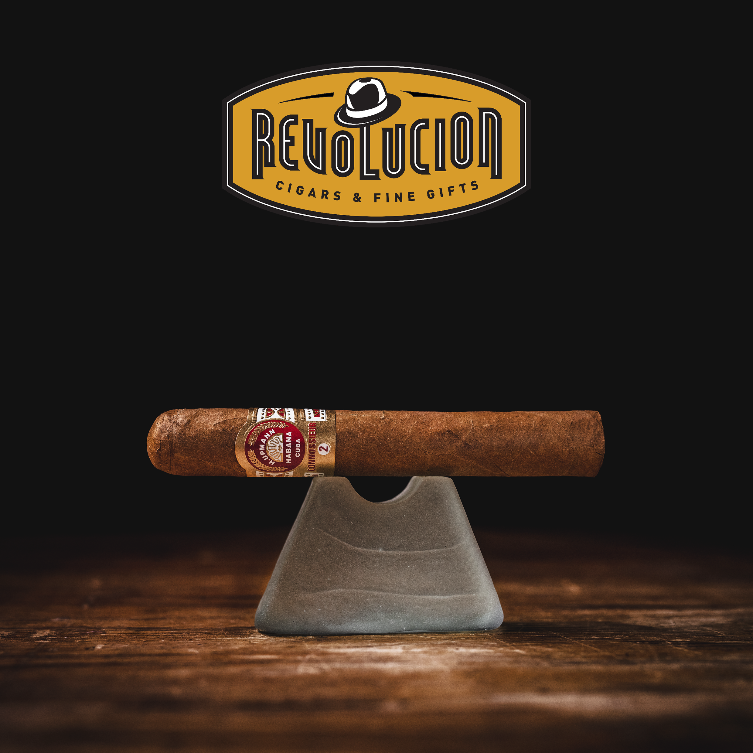 H. Upmann Connossieur No. 2 Cuban cigar resting on a glass stand with the Revolucion Cigars & Fine Gifts logo in the background.