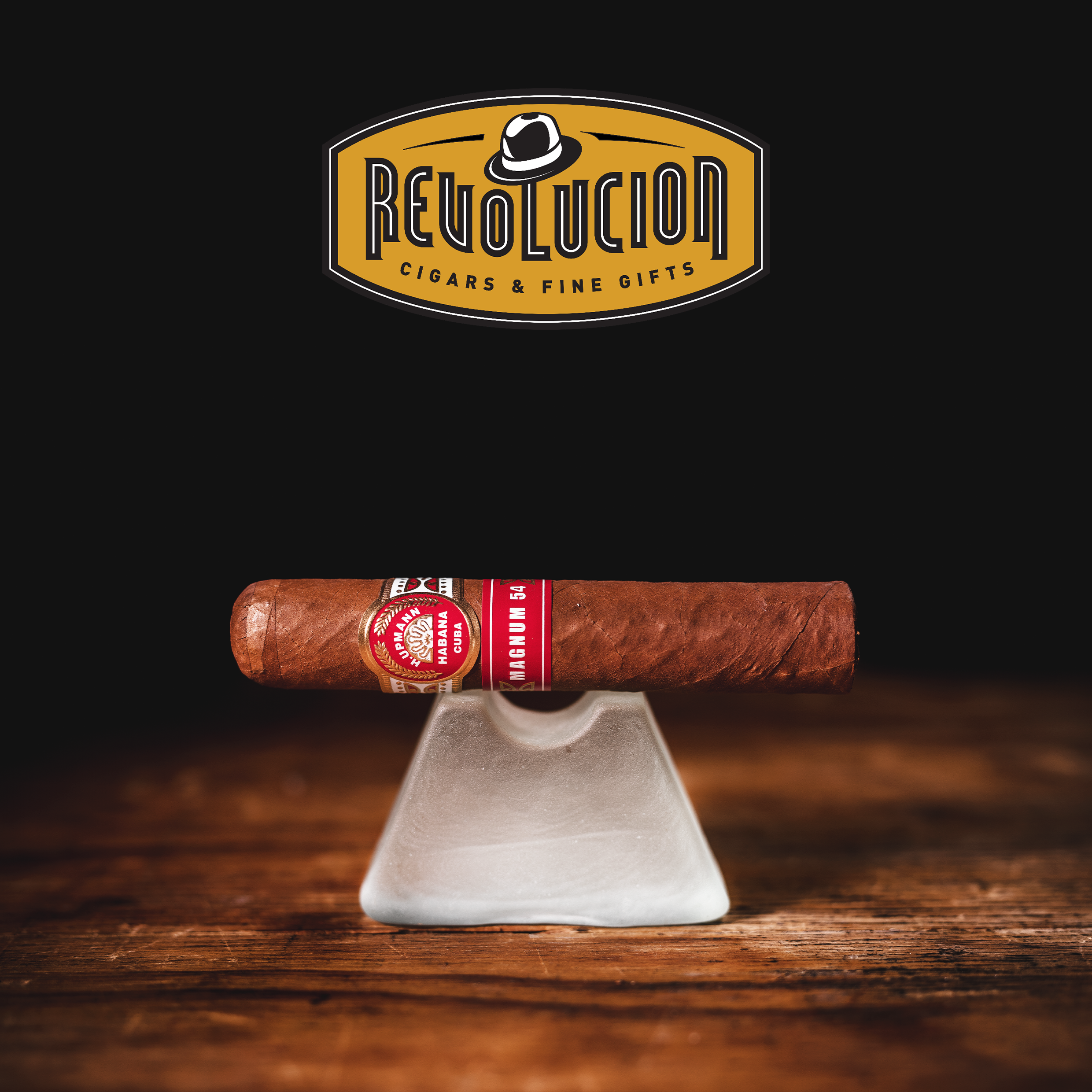 H. Upmann Magnum 54 Cuban cigar resting straightlly on a frosted glass stand, with the Revolucion Cigars & Fine Gifts logo above, set against a dark background.