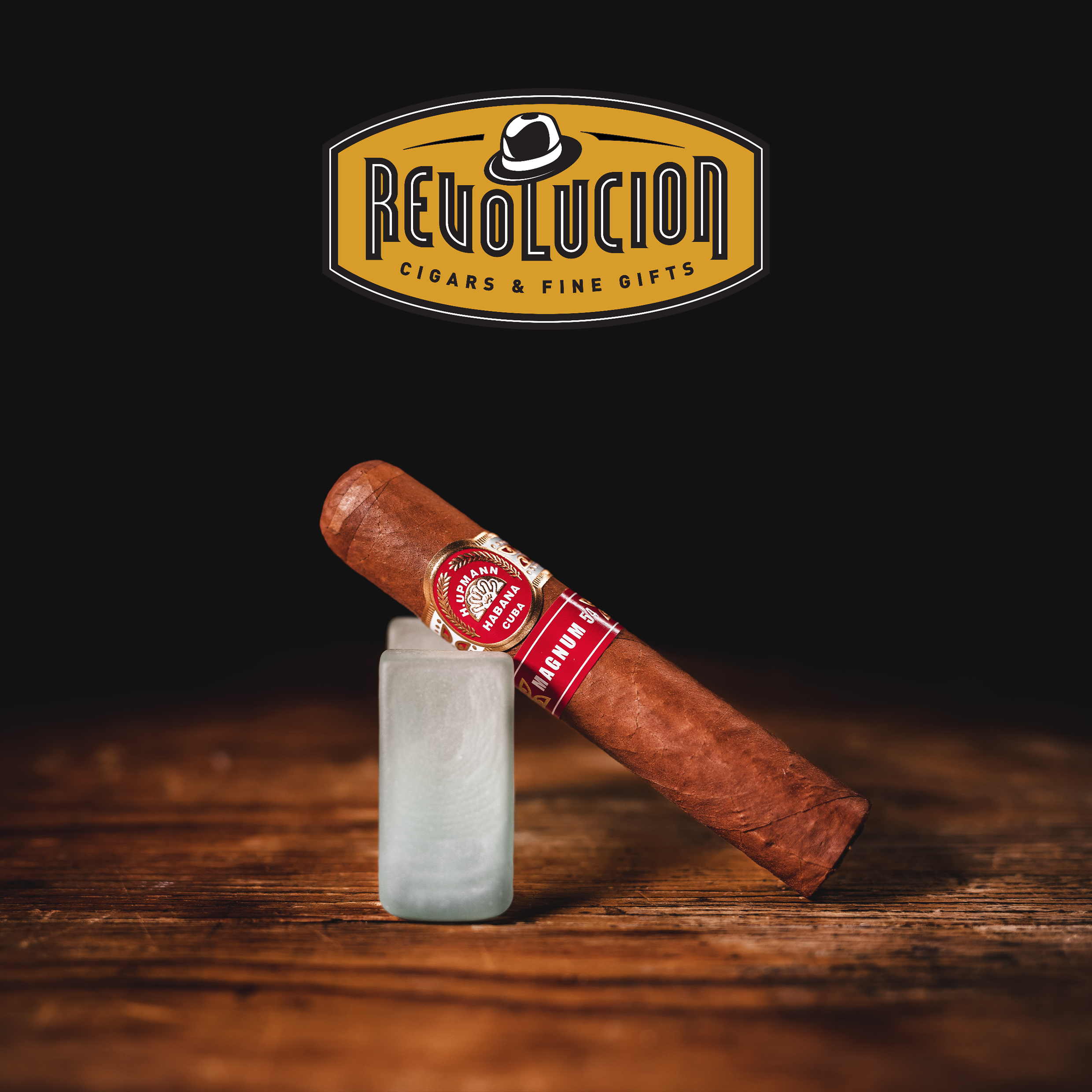 H. Upmann Magnum 54 Cuban cigar resting diagonally on a frosted glass stand, with the Revolucion Cigars & Fine Gifts logo above, set against a dark background.