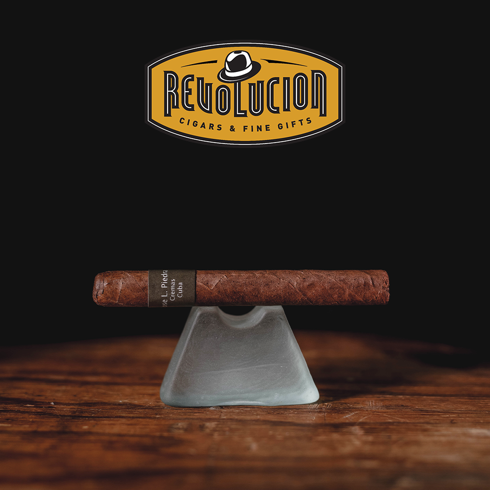 Jose L. Piedra Cremas Cuban cigar resting on a wooden surface with the Revolucion Cigars & Fine Gifts logo in the background.