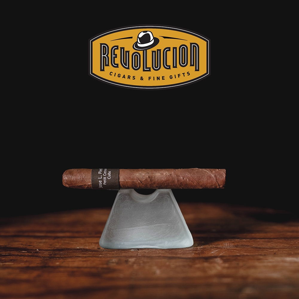 José L. Piedra Petit Cetros medium-strength Cuban cigar resting on a frosted glass, displayed on a wooden surface with a black background.
