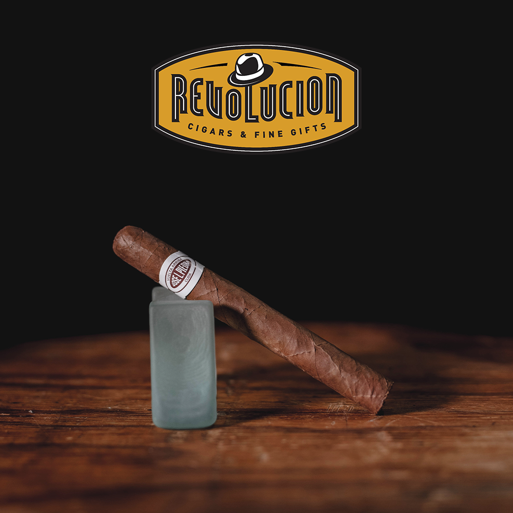 José L. Piedra Petit Cetros medium-strength Cuban cigar resting on a frosted glass, displayed on a wooden surface with a black background.