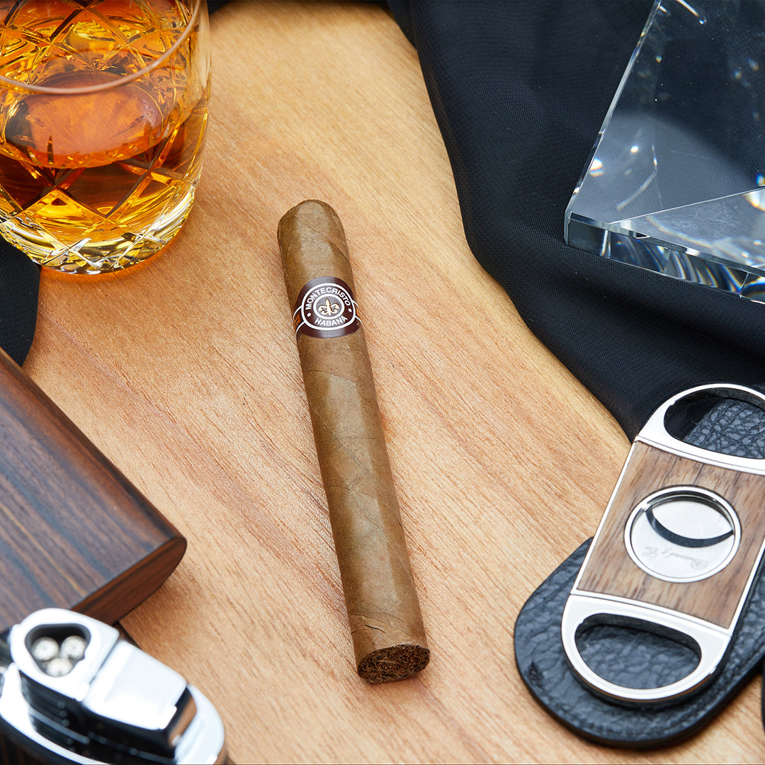 Montecristo No.4 Cuban cigar with a brown and white band, resting on a wooden surface alongside a cigar cutter, lighter, and a glass of whiskey.