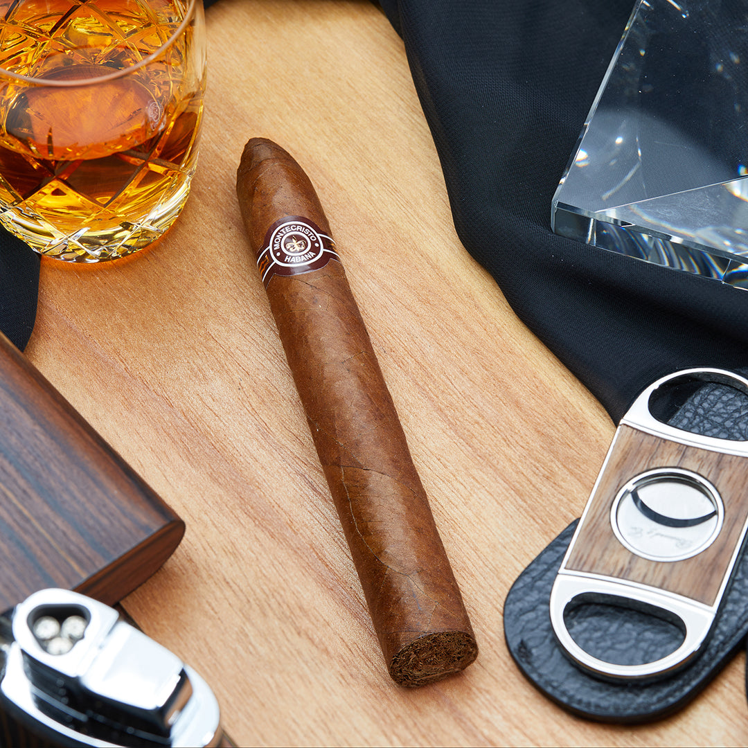 Montecristo No.2 Cuban torpedo cigar with a brown and white band, placed on a wooden surface alongside a cigar cutter, lighter, and a glass of whiskey.