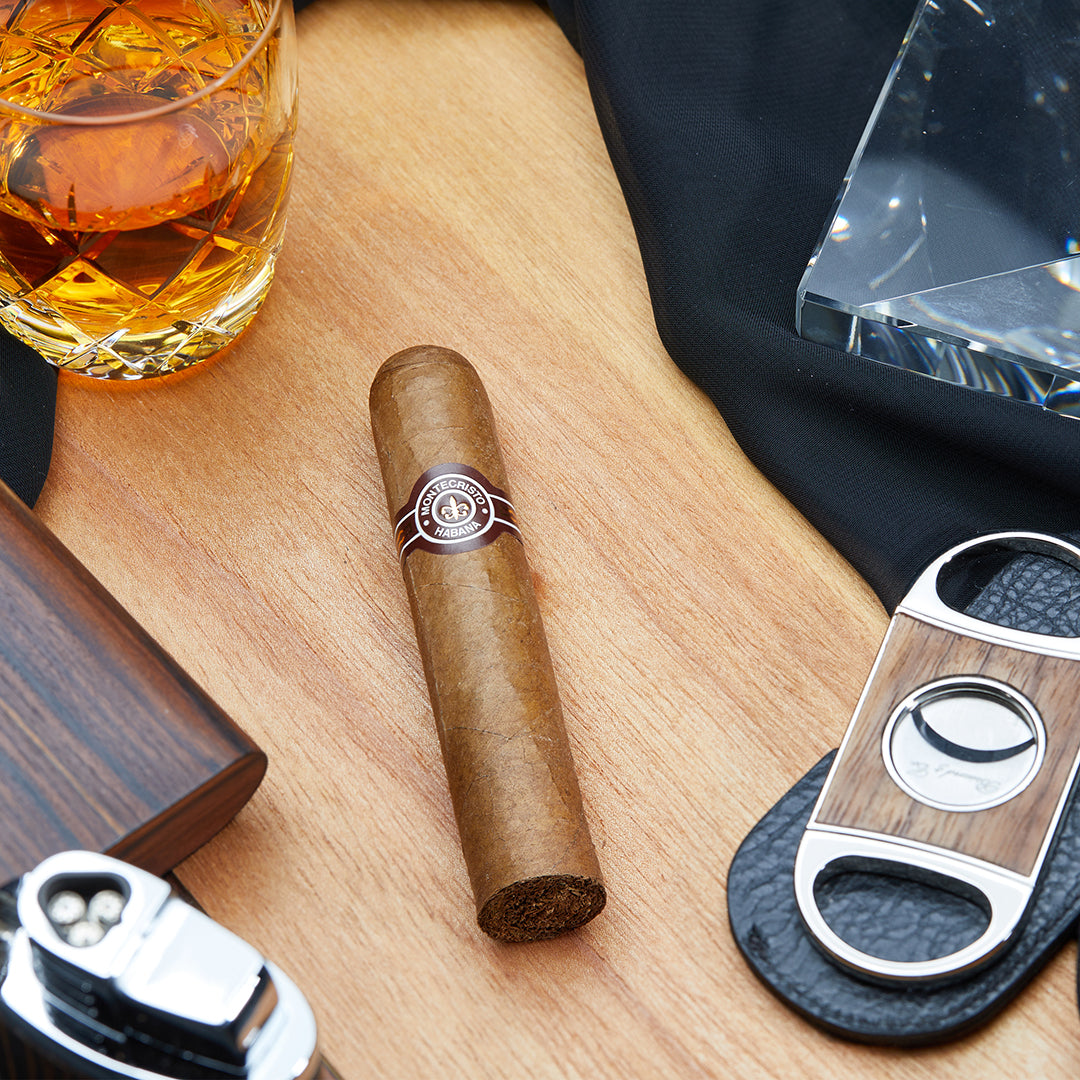 Montecristo Petit Edmundo Cuban cigar with a brown and white band, placed on a wooden surface with a cigar cutter, lighter, and a glass of whiskey.